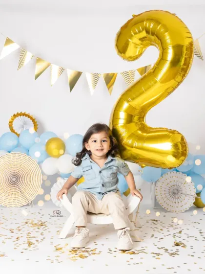A series of portraits celebrating a second birthday. The toddler poses with a giant gold '2' balloon, surrounded by blue and white balloons and confetti for a festive look.