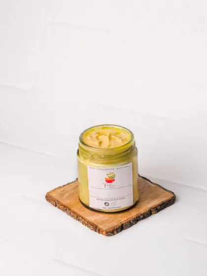 A jar of artisanal garlic hummus or a similar creamy dip, presented on a wooden coaster. The white background ensures the product and its label are the main focus.