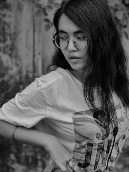 This black and white fashion portrait shows a young woman in a casual t-shirt and glasses, looking over her shoulder. The natural, outdoor setting and her relaxed pose give this a candid, lifestyle feel, proving that style is about attitude, not just clothes.