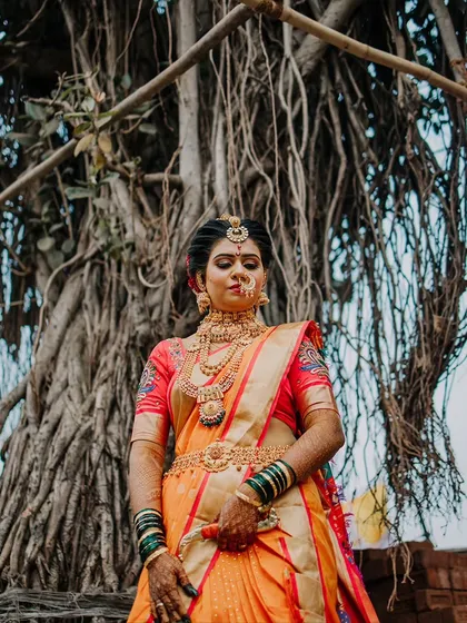 A powerful portrait of a Maharashtrian bride in a stunning orange saree, standing before a large banyan tree.