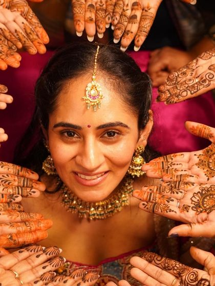A creative and beautiful shot focusing on the bride's face surrounded by mehendi-adorned hands. It's a classic photo that celebrates the artistry and community of the mehendi ceremony.