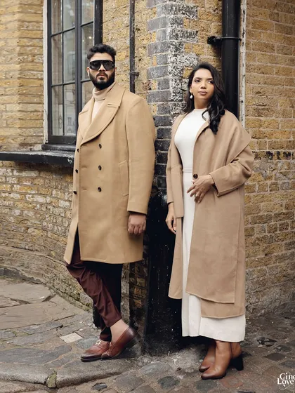 Posing against a rustic brick wall in London. The contrast between his burgundy trousers and their beige coats adds a subtle pop of color to the otherwise neutral palette.