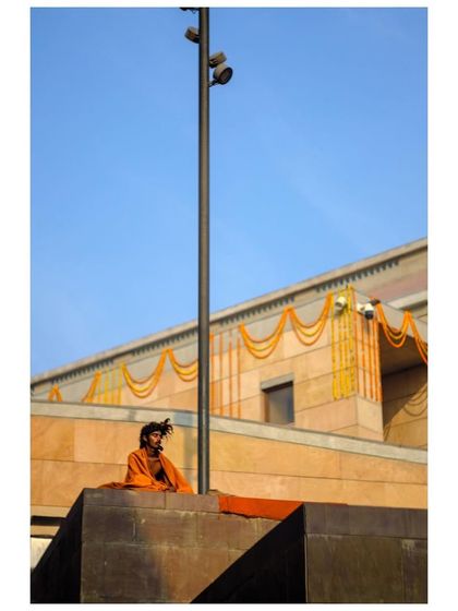A sadhu in orange robes sits meditating against a modern building in Varanasi. The vibrant colors contrast with the stark geometry of the architecture.