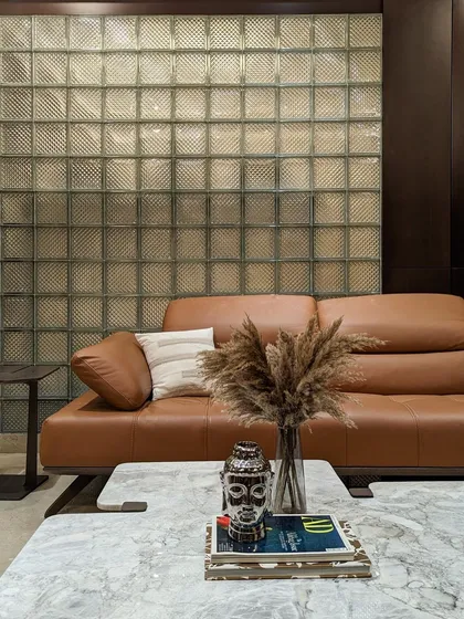 The family lounge features a full-height glass block wall, which acts as a textured, translucent screen for the mandir behind it. A comfortable brown leather sofa and marble coffee table complete this cozy, contemporary space.