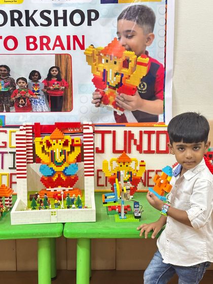 A young participant stands beside a table displaying multiple LEGO Ganesha creations from the workshop, showing the collective creativity of the class.