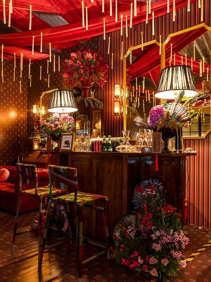 A wide shot of the bar area, where floating candles are reflected in mirrors behind the bar. The space is filled with lush floral arrangements, elegant lamps, and custom furniture, creating a layered and sophisticated lounge.