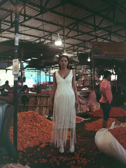 The model stands in a white fringe dress amidst piles of marigolds. The minimalist styling of the dress creates a stark, modern contrast with the vibrant, chaotic, and traditional setting of the flower market.