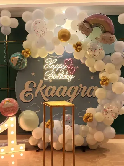 A whimsical rainbow and cloud-themed first birthday. The setup includes a custom backdrop, a pastel balloon arch, and a marquee number '1'.