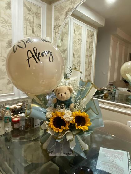 A cheerful and elegant welcome for baby Akay. This bouquet combines a personalized "Welcome Akay" balloon with a sweet teddy bear and bright, sunny sunflowers, creating a gift that is full of warmth and joy.