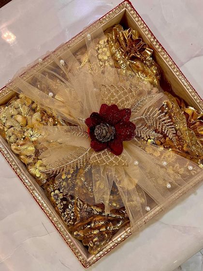 A beautifully decorated tray filled with dry fruits and sweets for a 'shagun' ceremony. The packing is done with gold net, intricate ribbons, and a floral centerpiece to match the festive wedding theme.