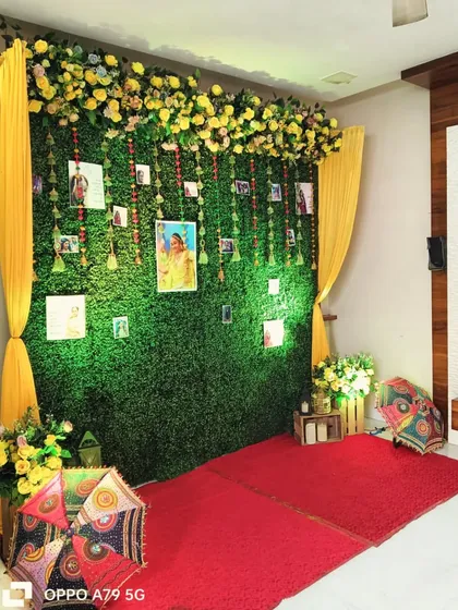 A lovely home Haldi decoration. I created a photo wall using a green foliage backdrop, hanging pictures of the family, and bright yellow drapes for a pop of color.