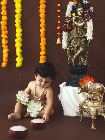 A simple and pure portrait of a baby dressed as Krishna, sitting beside a statue of the deity and playing with lotus flowers.