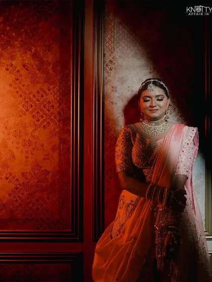 A stunning bridal portrait using dramatic lighting. The bride, in her delicate pink lehenga, is illuminated by a single spotlight against a rich, textured wall, creating an artistic and glamorous image.