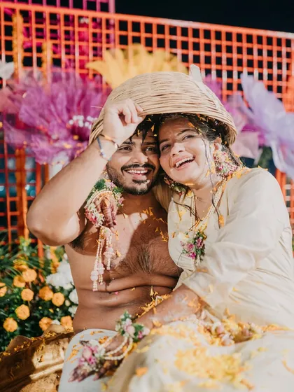 A fun and playful Haldi moment. The couple shares a laugh under a basket, surrounded by a colorful backdrop of giant paper flowers and a vibrant orange lattice screen.
