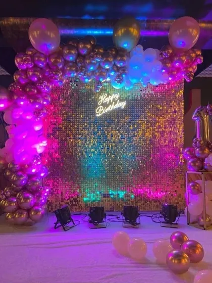 A vibrant party backdrop with a gold sequin wall lit by colorful party lights. A balloon arch and neon sign complete this fun and flashy setup for a first birthday.