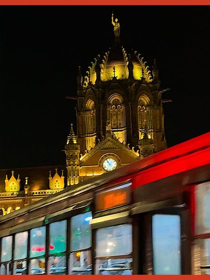 A motion-blurred shot of a red bus passing the magnificent CSMT station, its dome lit up against the night sky.
