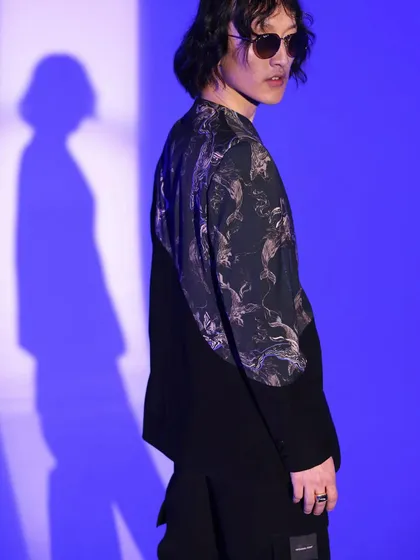 This shot from a menswear lookbook features a jacket with a graphic print panel against a vibrant blue background. The styling is edgy and modern, highlighting the unique design details of the garment.