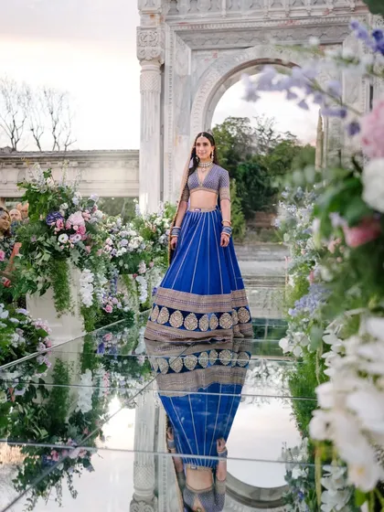 A bride in a stunning royal blue lehenga walks down a mirrored aisle lined with lush flowers at her destination wedding in Turkey. Her reflection creates a breathtaking visual, a moment of pure elegance we meticulously planned.