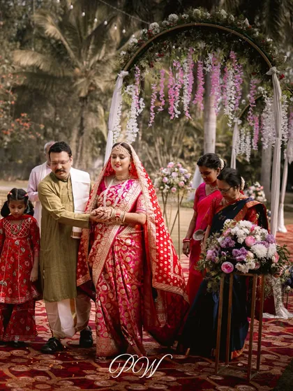 The bride's grand entrance, walking down an aisle flanked by family and beautiful floral arrangements. The path leads to a dreamy mandap with hanging wisteria, setting the stage for a magical ceremony.