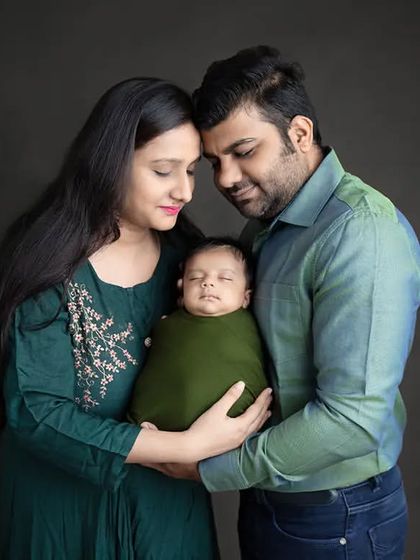 A studio portrait of a mother and father holding their newborn baby, wrapped in a green swaddle.