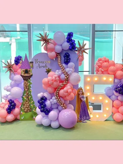 A fairytale come to life for a 5th birthday with our Rapunzel theme. This setup includes a large light-up number '5', a custom backdrop with Princess Avah's name, a Rapunzel tower cutout, and a flowing balloon garland in shades of purple and pink with golden starburst accents.