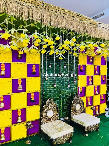 A unique Haldi backdrop with a checkerboard design using yellow marigolds and purple flowers. The setup includes traditional seating and is accented with hanging bells for a festive touch.