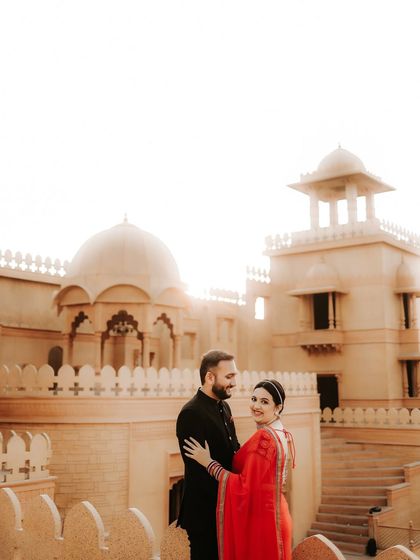 A classic couple portrait set against a magnificent heritage building. Their formal black and vibrant red outfits create a striking contrast with the sun-kissed stone, embodying royal elegance.