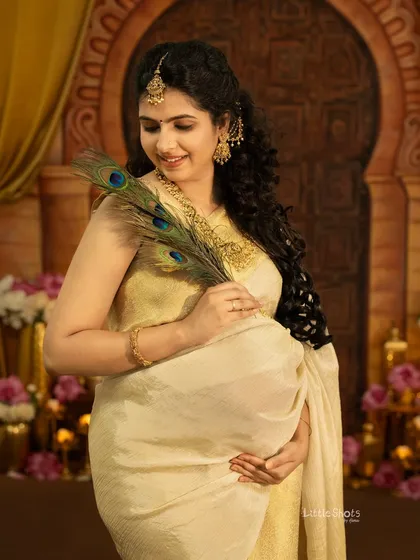 A beautiful portrait of an expecting mother in a traditional cream and gold saree, holding peacock feathers. She is standing in a regal setting, looking down at her bump with a gentle smile.