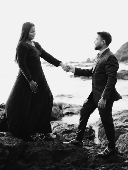 A full-body shot in black and white, capturing the couple's interaction on a rocky beach. The contrast between their dark outfits and the bright background is visually striking.