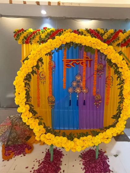A colorful Haldi setup with a circular marigold backdrop. The decor includes a small stool for the ceremony, Rajasthani umbrellas, and a floor scattered with rose petals.