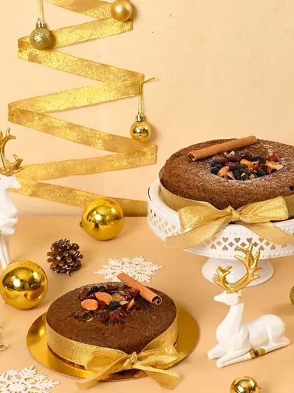 Two traditional Christmas fruitcakes decorated with gold ribbons and surrounded by festive ornaments. The warm, golden tones create an elegant and classic holiday mood.