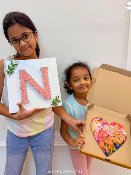 Sisters showcasing their different art projects. One created a personalized string art initial, while the other made a vibrant fluid art heart.