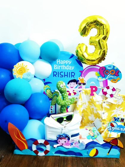 A pool party themed hamper is perfect for a summer birthday. This gift for a 3-year-old includes a fun shirt, themed decorations like a surfboard and lifebuoy, and blue balloons to create a splash.