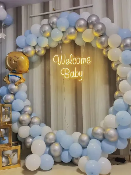 A stylish welcome home setup for a baby boy. This design features a full ring of blue, white, and silver balloons, a "Welcome Baby" neon sign, and "BABY" blocks filled with matching balloons.