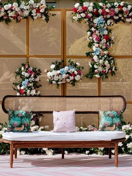 The main seating stage for the pastel Mehendi, featuring a modern rattan bench against a cane-paneled backdrop adorned with asymmetrical floral arrangements.
