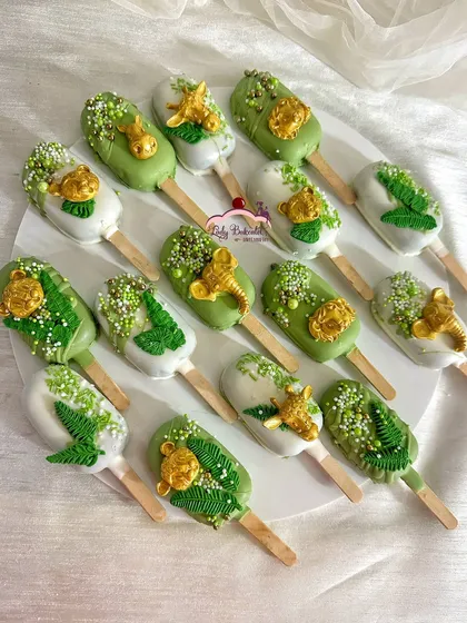 A close-up of my jungle-themed cakesicles. I use a mix of green and white chocolate coatings, detailed with edible gold animal figures, delicate leaf patterns, and sprinkles to make each one a tiny work of art.