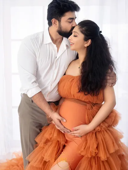 A tender moment captured against a bright, airy background. The partner kisses his wife's forehead as they embrace, both focused on the baby bump.