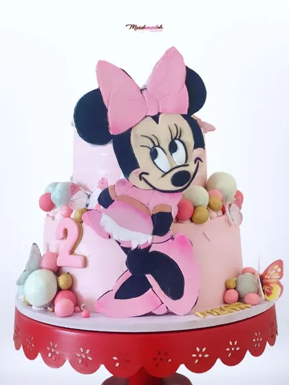A grand two-tiered Minnie Mouse cake for a second birthday. The highlight is the large, 10-inch hand-cut 2D fondant Minnie, surrounded by colourful cake pops and edible butterflies.