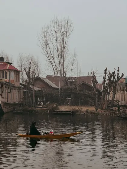A lone shikara glides through the water, with the rustic wooden houseboats and bare winter trees creating a moody, atmospheric backdrop.