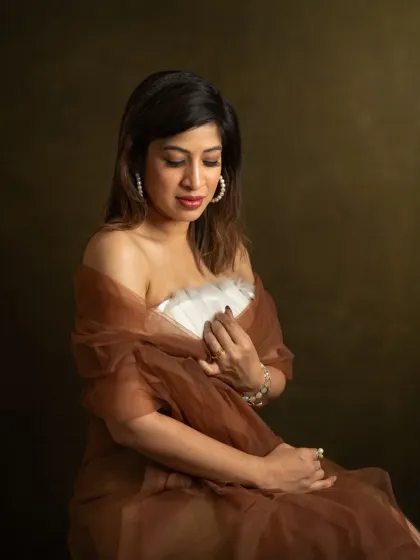 A moment of quiet contemplation. Pallavi, holding a traditional fan, looks down with a gentle expression. The warm, focused light and rich brown tones give this portrait a painterly quality, emphasizing emotion and texture over everything else.