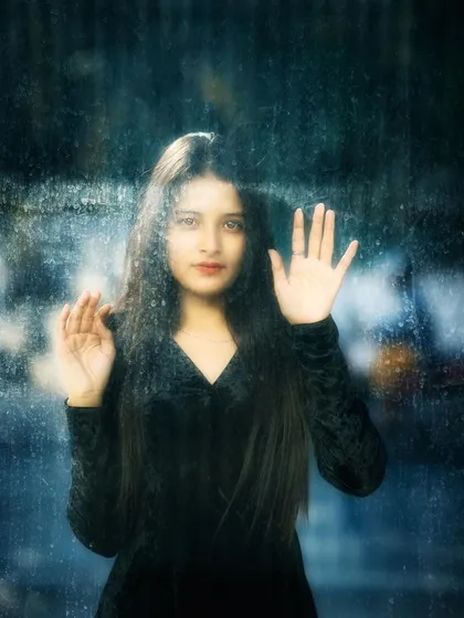 A creative and artistic portrait of a woman seen through a rain-streaked window. Her hand rests on the glass, creating a sense of longing and painterly beauty, perfect for a conceptual photoshoot.
