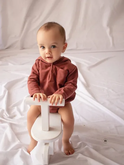 A simple and classic sitter portrait. A baby in a rust-colored romper sits on a small white toy bike against a clean backdrop.