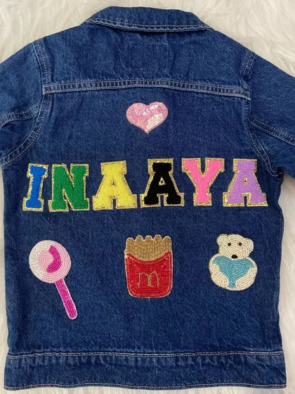A fun and stylish custom denim jacket for 'Inaaya'. Her name is spelled in multi-colored glittery letters, surrounded by beaded patches of a lollipop, french fries, and a teddy bear.