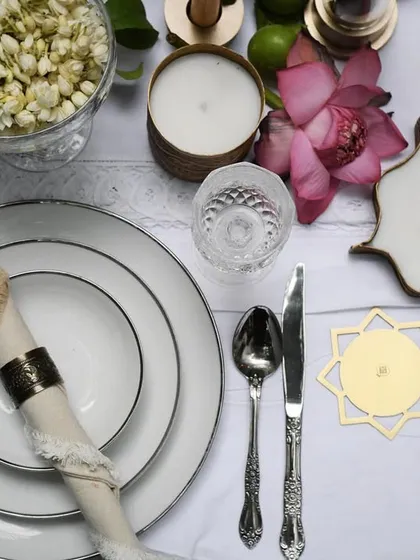 An overhead flat lay of a festive table setting for Byaas Home. This detailed shot highlights the beautiful interplay between the crisp white dinnerware, intricate silverware, and the brand's unique star-shaped candles and coasters.