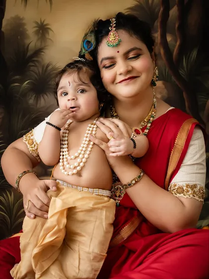 A tender moment between mother and child. The baby, dressed as Krishna, curiously tastes his fingers while his mother holds him close.