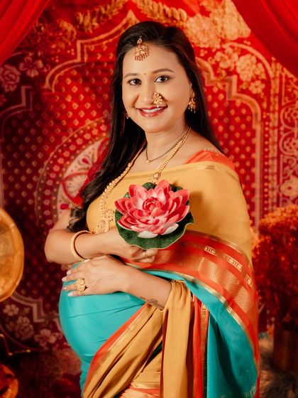 A smiling portrait of a mom-to-be in a colorful saree, holding a lotus flower. The rich red background and traditional jewelry complete this vibrant and happy maternity photo.