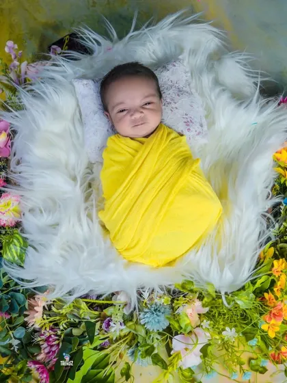 An awake newborn wrapped in yellow, looking bright-eyed in a nest of fur and flowers.