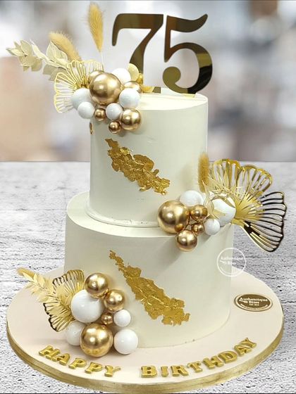 A stylish two-tiered cake celebrating a 75th birthday. The design uses white and gold spheres and dried palm leaves for a modern and sophisticated milestone cake.