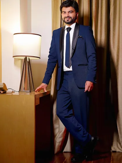 A sophisticated portrait of the groom in a sharp blue suit, leaning against a desk. The warm lamp light adds a cozy and intimate feel.