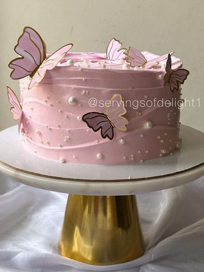 This pink butterfly cake is displayed on a beautiful marble and gold stand, highlighting its elegance. It's perfect for birthdays or bridal showers.